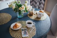 Round breakfast table with white flowers, golden mats, coffee, croissant and smartphone