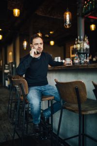 Handsome young man with smartphone drinking whisky at bar or pub