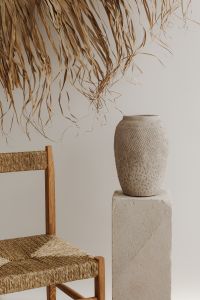 Kaboompics - A textured clay vase placed on a minimal stone pedestal next to a wooden chair with woven seating - creating a natural - earthy aesthetic