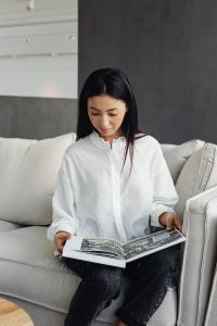 A young Asian woman sits on the couch and reads a book or magazine