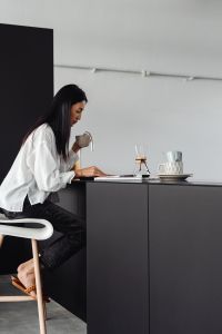 Adult young Asian woman sits on stool in kitchen - drinks coffee - reads magazine