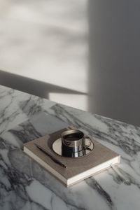 Kaboompics - Chrome Espresso Cup on a Closed Beige Notebook, Marble Workspace with Strong Window Shadows