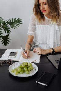 Kaboompics - Businesswoman Writing in Notebook Beside Smartphone and Fruit at Office Desk