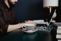 Kaboompics - Man Typing on Laptop with Cappuccino, Earbuds and Stacked Books in Foreground