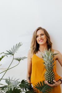 Kaboompics - Smiling Woman Holding Pineapple at Her Side with Green Leaves in Foreground