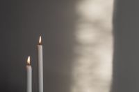 Kaboompics - Two Lit White Taper Candles Against a Soft Gray Wall with Sunlit Shadows, Minimal Home Atmosphere
