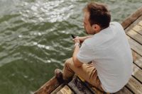 Kaboompics - Man Sitting on Wooden Lakeside Dock Using Smartphone, Casual Outdoor Lifestyle Photography