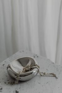 Kaboompics - Silver Chain Draped Across Sculptural Chrome Bowl on Speckled Stone Table, Minimal Jewelry Still Life