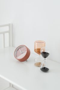 Kaboompics - Vertical Still Life of a Rose-Pink Clock with Amber and Black Hourglasses on White Table