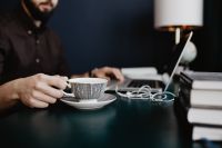 Kaboompics - Male Designer Holding Coffee while Working on Laptop beside Stacked Books