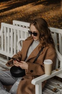 Kaboompics - Woman in Sunglasses Using Phone with Coffee on a White Park Bench