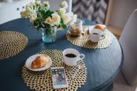 Kaboompics - Round breakfast table with white flowers, golden mats, coffee, croissant and smartphone
