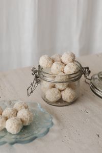 Kaboompics - Open Glass Jar of Coconut Truffles with White Plate on Textured Stone, Bright Home Snack Still Life