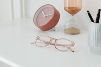 Kaboompics - Rose-Gold Eyeglasses in Foreground with Soft-Focus Pink Clock, Amber Hourglass and Pen Cup