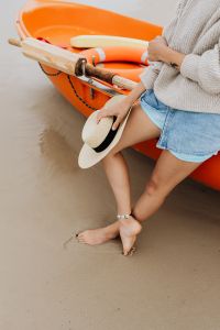 Kaboompics - Woman Holding a Straw Sun Hat While Sitting Beside an Orange Kayak, Barefoot Beach Portrait