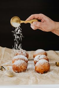 Kaboompics - Hand Sprinkling Powdered Sugar onto Polish Paczki with a Gold Spoon, Dark Backdrop