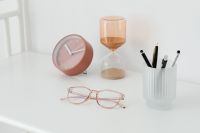 Kaboompics - Rose-Gold Eyeglasses with Pink Clock, Amber Hourglass and Pens in a White Desk Still Life