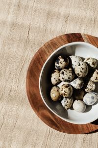Kaboompics - Speckled Eggs in a White Bowl on a Wooden Board, Linen Copy Space for Easter Recipes