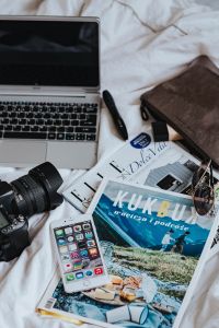 Silver laptop, a camera, magazines and other items on white bed sheets