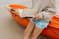 Kaboompics - Woman Reading an Open Book While Sitting in an Orange Kayak on the Sandy Shore