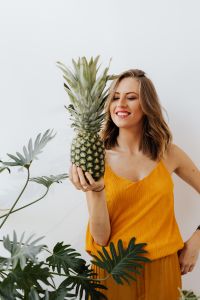 Kaboompics - Woman Looking at a Pineapple beside Her Face in a Yellow Top, Fresh Lifestyle Portrait