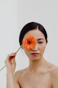 Kaboompics - Asian Beauty Portrait Holding Orange Gerbera Over One Eye, Purple Eyebrow Makeup and Copy Space