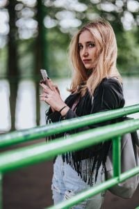 Kaboompics - Blonde woman in a black jacket and ripped jeans by a green handrail