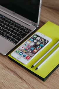 Silver Acer Laptop, White Apple iPhone And Notepad Workspace On Wooden Desk