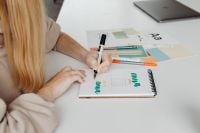 Kaboompics - Graphic Designer Sketching Lettering on a Spiral Notebook beside Color Swatches, Markers and Laptop