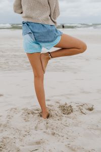 Kaboompics - Rear View of a Woman Practicing Tree Pose on the Beach in Sweater and Denim Shorts