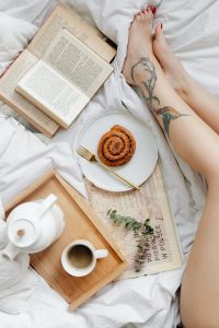 Kaboompics - Cinnamon Roll and Paperback beside a Woman’s Tattooed Legs on White Bedding