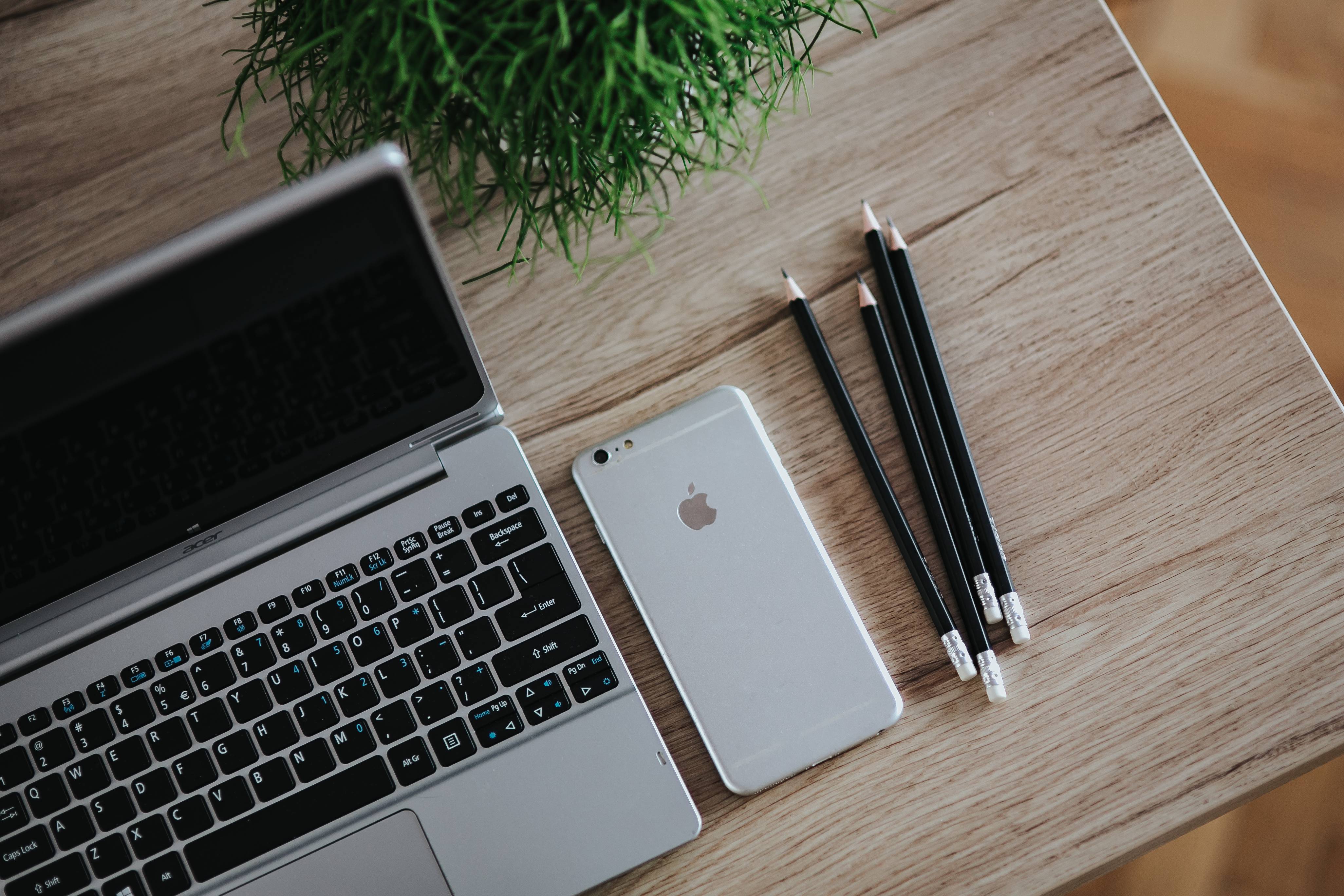 Silver Acer laptop on a wooden desk with a green plant, pencils and an Apple iPhone